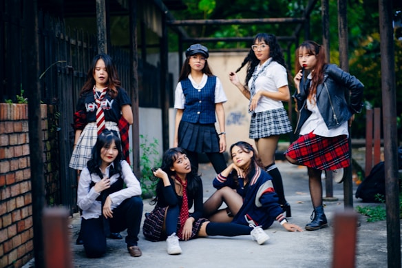 A group of seven young women are gathered together outdoors, posing confidently. They are dressed in a variety of schoolgirl-inspired outfits with plaid skirts, blouses, and some casual wear. The setting appears to be an alleyway or courtyard with a mixture of urban and natural elements. Some are standing while others are sitting or kneeling, giving the scene a dynamic feel.