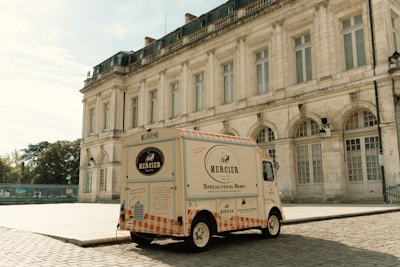 A vintage-style delivery van is parked on a cobblestone street in front of a grand, historic building. The van features branding for 'Mercier Specialités du Berry,' indicating it offers artisan specialties from the Berry region. The building has large arched windows and classical architectural details, contributing to an old-world charm. Soft lighting suggests it's either early morning or late afternoon.