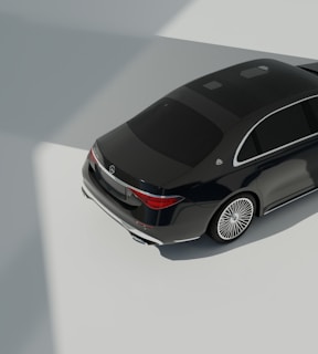 A dark luxury car is parked on a smooth, light-colored surface with clear reflections. The car features elegant design elements such as sleek curves and shiny metallic accents. The lighting creates strong shadows and highlights, suggesting an indoor environment with natural light coming from above.