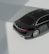 A dark luxury car is parked on a smooth, light-colored surface with clear reflections. The car features elegant design elements such as sleek curves and shiny metallic accents. The lighting creates strong shadows and highlights, suggesting an indoor environment with natural light coming from above.
