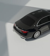 A dark luxury car is parked on a smooth, light-colored surface with clear reflections. The car features elegant design elements such as sleek curves and shiny metallic accents. The lighting creates strong shadows and highlights, suggesting an indoor environment with natural light coming from above.