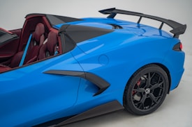 A sleek, blue sports car showcasing its aerodynamic design with a prominent rear spoiler, black rims, and a vibrant red interior. The image emphasizes the car&rsquo;s modern and sporty aesthetic.