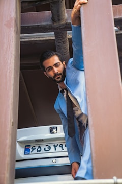 A man wearing a light blue suit and black tie is posing among large metal beams. He appears to be crouching or leaning, with one hand gripping a beam above him. The background includes a vehicle with an Iranian license plate.
