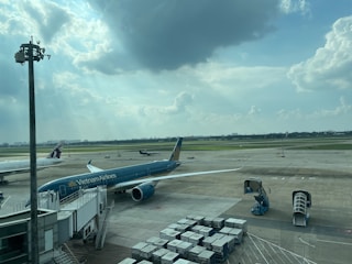 A busy airport scene with a Vietnam Airlines airplane connected to a jet bridge. Another aircraft is seen taxiing on the runway in the background, while various ground support equipment and cargo containers are present. The sky is partly cloudy with sunlight streaming through the clouds.