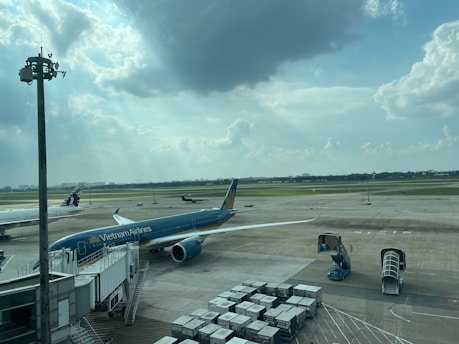 A busy airport scene with a Vietnam Airlines airplane connected to a jet bridge. Another aircraft is seen taxiing on the runway in the background, while various ground support equipment and cargo containers are present. The sky is partly cloudy with sunlight streaming through the clouds.