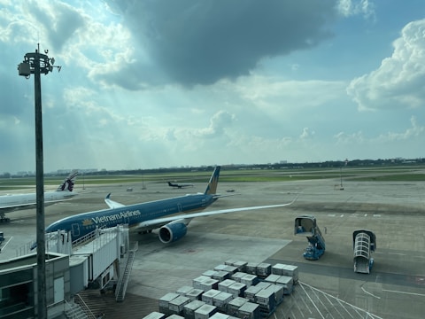 A busy airport scene with a Vietnam Airlines airplane connected to a jet bridge. Another aircraft is seen taxiing on the runway in the background, while various ground support equipment and cargo containers are present. The sky is partly cloudy with sunlight streaming through the clouds.