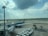 A busy airport scene with a Vietnam Airlines airplane connected to a jet bridge. Another aircraft is seen taxiing on the runway in the background, while various ground support equipment and cargo containers are present. The sky is partly cloudy with sunlight streaming through the clouds.