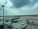 A busy airport scene with a Vietnam Airlines airplane connected to a jet bridge. Another aircraft is seen taxiing on the runway in the background, while various ground support equipment and cargo containers are present. The sky is partly cloudy with sunlight streaming through the clouds.