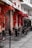 A charming outdoor café setting with several metal tables and chairs arranged neatly on a tiled pavement. Vibrant red flowers cascade down the building facade, creating a striking contrast with the muted pastel tones of the walls. The ambiance is quiet and inviting, suggesting a peaceful spot for a meal or coffee.