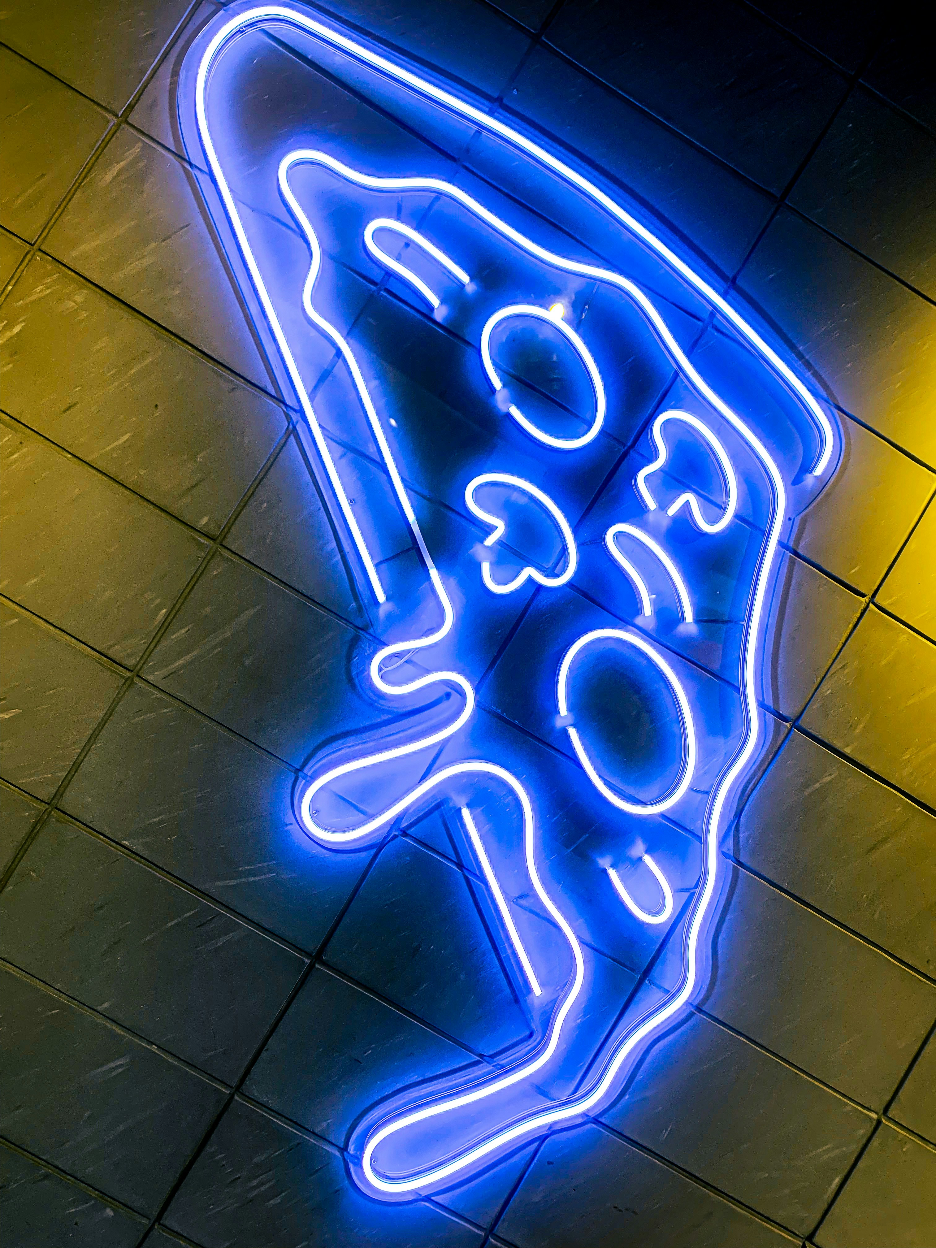 A neon sign depicting a slice of pizza is mounted on a tiled wall. The neon tubing is bright blue and highlights the shape of a pizza slice with details like pepperoni and mushrooms.