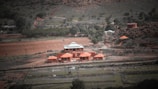 A rural landscape features a compound of red-roofed buildings amidst greenery and hills. The scene includes a winding dirt road with scattered trees and bushes, suggesting a natural yet cultivated environment. There appear to be livestock grazing in the open fields surrounding the structures.
