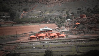 A rural landscape features a compound of red-roofed buildings amidst greenery and hills. The scene includes a winding dirt road with scattered trees and bushes, suggesting a natural yet cultivated environment. There appear to be livestock grazing in the open fields surrounding the structures.