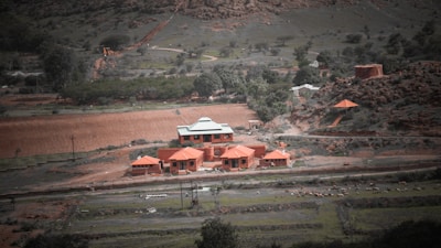 A rural landscape features a compound of red-roofed buildings amidst greenery and hills. The scene includes a winding dirt road with scattered trees and bushes, suggesting a natural yet cultivated environment. There appear to be livestock grazing in the open fields surrounding the structures.