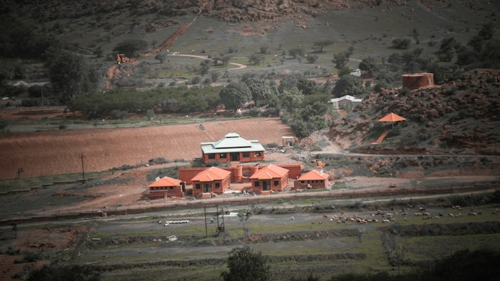 A rural landscape features a compound of red-roofed buildings amidst greenery and hills. The scene includes a winding dirt road with scattered trees and bushes, suggesting a natural yet cultivated environment. There appear to be livestock grazing in the open fields surrounding the structures.