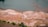 An aerial view of a landscape with a large body of water at the top, transitioning to an expansive area of land. The land has patches of green and orange tones, suggesting a mixture of grass and bare soil. There are a few scattered trees and what appear to be animals or people, small in the scale of the image, moving across the landscape.