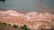 An aerial view of a landscape with a large body of water at the top, transitioning to an expansive area of land. The land has patches of green and orange tones, suggesting a mixture of grass and bare soil. There are a few scattered trees and what appear to be animals or people, small in the scale of the image, moving across the landscape.