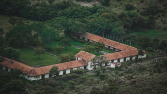 An aerial view of a building complex with red-tiled roofs forming a 'U' shape amidst green trees and vegetation. The building appears to be surrounded by a lush, forested area with a mix of open spaces and dense tree cover.