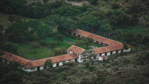 An aerial view of a building complex with red-tiled roofs forming a 'U' shape amidst green trees and vegetation. The building appears to be surrounded by a lush, forested area with a mix of open spaces and dense tree cover.