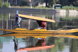 A traditional wooden boat with a yellow canopy floats on calm water, reflecting its image. The boat is ornately decorated with blue fabric and features a maroon curtain. In the background, a stone wall and lush greenery provide a serene setting.