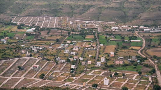 An aerial view of a semi-urban area with planned plots. There are scattered houses, green agricultural fields, and winding roads. Surrounding the development are hills with sparse vegetation.