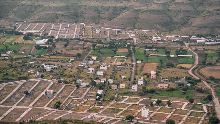 An aerial view of a semi-urban area with planned plots. There are scattered houses, green agricultural fields, and winding roads. Surrounding the development are hills with sparse vegetation.