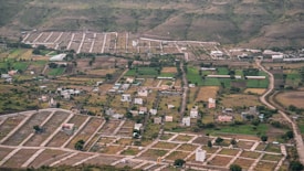 An aerial view of a semi-urban area with planned plots. There are scattered houses, green agricultural fields, and winding roads. Surrounding the development are hills with sparse vegetation.