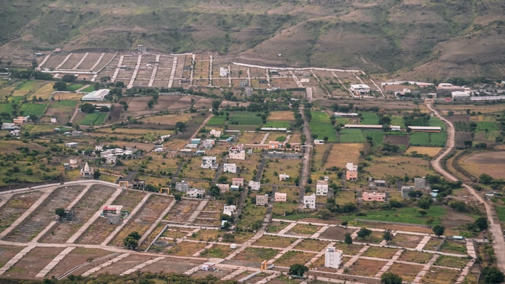 An aerial view of a semi-urban area with planned plots. There are scattered houses, green agricultural fields, and winding roads. Surrounding the development are hills with sparse vegetation.