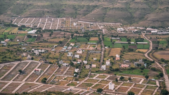 An aerial view of a semi-urban area with planned plots. There are scattered houses, green agricultural fields, and winding roads. Surrounding the development are hills with sparse vegetation.