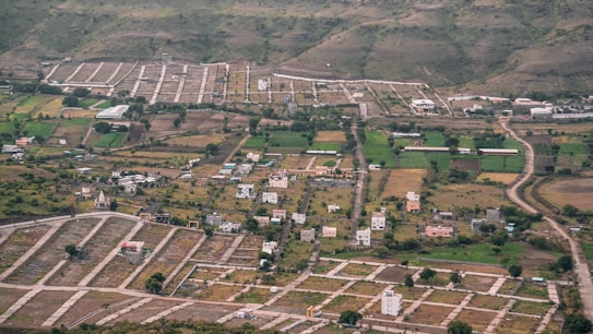 An aerial view of a semi-urban area with planned plots. There are scattered houses, green agricultural fields, and winding roads. Surrounding the development are hills with sparse vegetation.