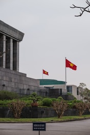 A large stone building with angular, modern architecture features a prominent Vietnamese flag waving on a tall flagpole. The surrounding area is landscaped with neatly trimmed bushes and trees. In the foreground, a sign in both Vietnamese and English reads 'DO NOT GO THIS WAY.' The sky appears overcast, adding a solemn ambiance.