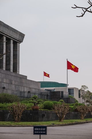 A large stone building with angular, modern architecture features a prominent Vietnamese flag waving on a tall flagpole. The surrounding area is landscaped with neatly trimmed bushes and trees. In the foreground, a sign in both Vietnamese and English reads 'DO NOT GO THIS WAY.' The sky appears overcast, adding a solemn ambiance.