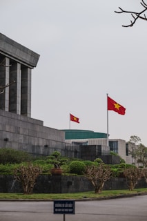 A large stone building with angular, modern architecture features a prominent Vietnamese flag waving on a tall flagpole. The surrounding area is landscaped with neatly trimmed bushes and trees. In the foreground, a sign in both Vietnamese and English reads 'DO NOT GO THIS WAY.' The sky appears overcast, adding a solemn ambiance.
