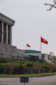 A large stone building with angular, modern architecture features a prominent Vietnamese flag waving on a tall flagpole. The surrounding area is landscaped with neatly trimmed bushes and trees. In the foreground, a sign in both Vietnamese and English reads 'DO NOT GO THIS WAY.' The sky appears overcast, adding a solemn ambiance.