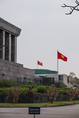 A large stone building with angular, modern architecture features a prominent Vietnamese flag waving on a tall flagpole. The surrounding area is landscaped with neatly trimmed bushes and trees. In the foreground, a sign in both Vietnamese and English reads 'DO NOT GO THIS WAY.' The sky appears overcast, adding a solemn ambiance.