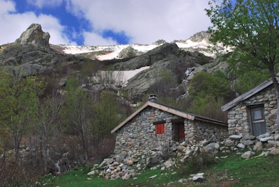 A rustic stone cabin with a wooden roof is nestled among rocky hills and lush greenery. Snow-capped peaks are visible in the background under a partly cloudy sky. Trees and bushes surround the cabin, contributing to the tranquil mountain setting.