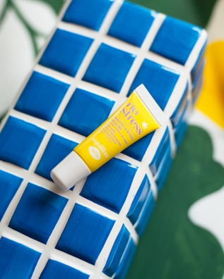 A yellow tube of lip balm labeled 'No Stress' rests diagonally on a blue and white ceramic, tiled platform. The background includes green and yellow shades, adding a vibrant and colorful contrast.