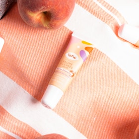 A tube of lip balm labeled 'BAUME LEVRES JOUR' with colorful graphics is placed on an orange and white striped fabric. There is a peach visible near the top left corner, suggesting a theme involving fruit or freshness.
