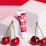 A tube of cherry pearly lip balm with a vibrant red and pink design stands against two white cube blocks. Three glossy red cherries with green stems are placed in front of the balm on a pink and red background.