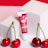 A tube of cherry pearly lip balm with a vibrant red and pink design stands against two white cube blocks. Three glossy red cherries with green stems are placed in front of the balm on a pink and red background.
