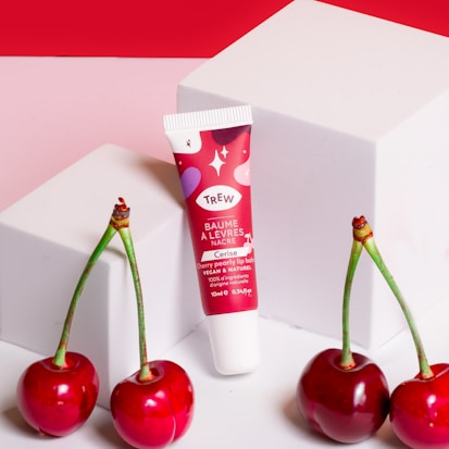 A tube of cherry pearly lip balm with a vibrant red and pink design stands against two white cube blocks. Three glossy red cherries with green stems are placed in front of the balm on a pink and red background.