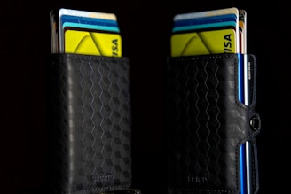 Two black wallets with a geometric hexagonal pattern are filled with colorful credit and bank cards sticking out. The wallets are positioned against a dark background, making the vivid colors of the cards—yellow, blue, and white—stand out prominently.