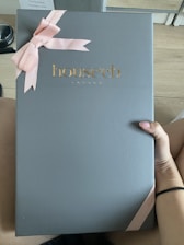 A large box with a sleek, matte gray surface features the embossed gold logo 'house cb London' at the center. A pink ribbon tied into a bow decorates the top left corner, adding an elegant touch. A hand is holding the box against a light wood floor background.