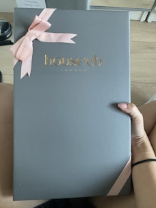 A large box with a sleek, matte gray surface features the embossed gold logo 'house cb London' at the center. A pink ribbon tied into a bow decorates the top left corner, adding an elegant touch. A hand is holding the box against a light wood floor background.