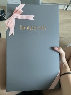 A large box with a sleek, matte gray surface features the embossed gold logo 'house cb London' at the center. A pink ribbon tied into a bow decorates the top left corner, adding an elegant touch. A hand is holding the box against a light wood floor background.