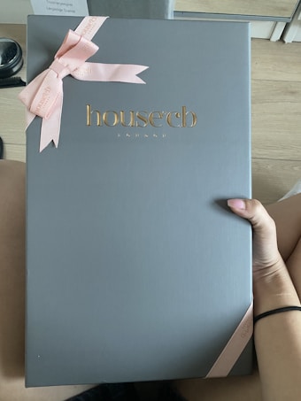 A large box with a sleek, matte gray surface features the embossed gold logo 'house cb London' at the center. A pink ribbon tied into a bow decorates the top left corner, adding an elegant touch. A hand is holding the box against a light wood floor background.