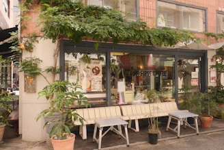A cozy outdoor café with a variety of potted plants and greenery framing the windows. The seating area includes a white bench and two small tables, creating an inviting atmosphere. Posters and signs can be seen in the windows, suggesting a lively and welcoming café environment.