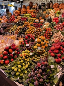 A vibrant market stall displays an array of colorful fruits, including pumpkins, apples, oranges, pineapples, and figs. The produce is neatly arranged in piles, creating a rich tapestry of colors and textures. Bright yellow bananas hang at the back, while small price tags are visible next to some items. The background features geometric tiles, adding a modern touch to the traditional setting.