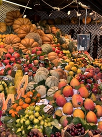 A vibrant and colorful market stall displaying a variety of fresh fruits. Large pumpkins and melons are prominently stacked at the back, while the front is filled with mangoes, papayas, apples, and other assorted fruits. The stall is well-lit and the fruits are neatly arranged, creating an eye-catching display.