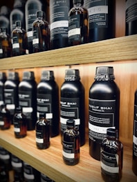 Multiple bottles of fragrance oil are neatly arranged on wooden shelves. The bottles vary in size and have labels with text both in English and another language. The lighting is warm, highlighting the sleek, dark-colored containers.