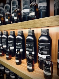 Multiple bottles of fragrance oil are neatly arranged on wooden shelves. The bottles vary in size and have labels with text both in English and another language. The lighting is warm, highlighting the sleek, dark-colored containers.